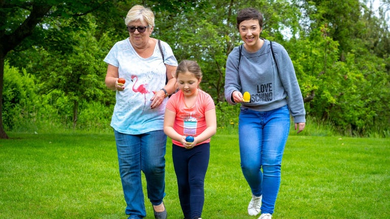 Two women and a girl are walking towards the camera trying to balance a wooden egg on a spoon in a race.
